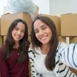 Two young women taking a selfie while moving, surrounded by cardboard boxes.